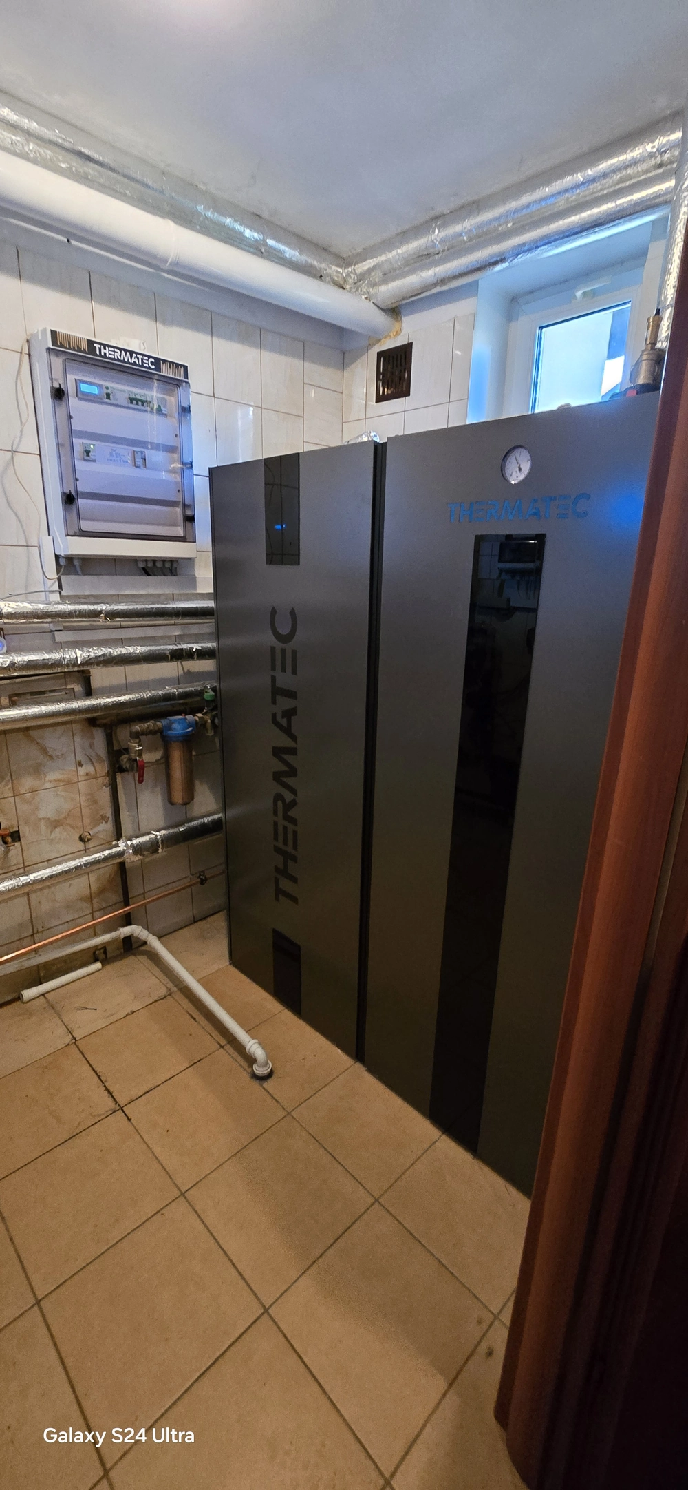 Czarna obudowa pompy ciepła powietrze woda wewnątrz kotłowni z widoczną instalacją rurową